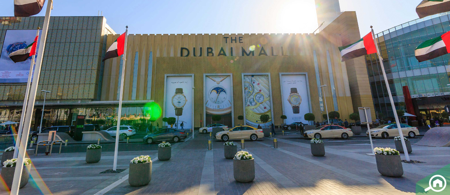 The image shows the entrance of The Dubai Mall, featuring large advertisements and flags, with cars and pedestrians in the foreground.